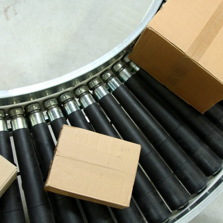 Three cardboard boxes on a curved conveyor belt with black rollers in an industrial setting.