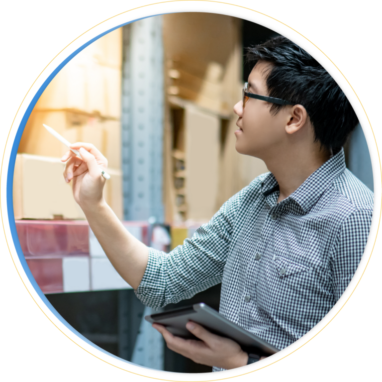 A person wearing glasses and a checkered shirt holds a tablet and points at boxes on warehouse shelves with a pen, focusing on inventory balancing.
