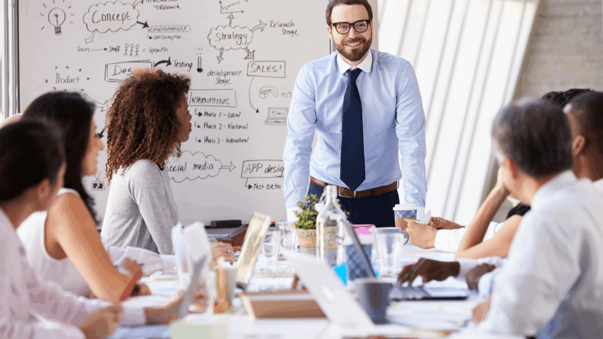 A man in business attire stands and smiles while presenting to a group of colleagues seated around a conference table, with a whiteboard full of notes and diagrams visible in the background.