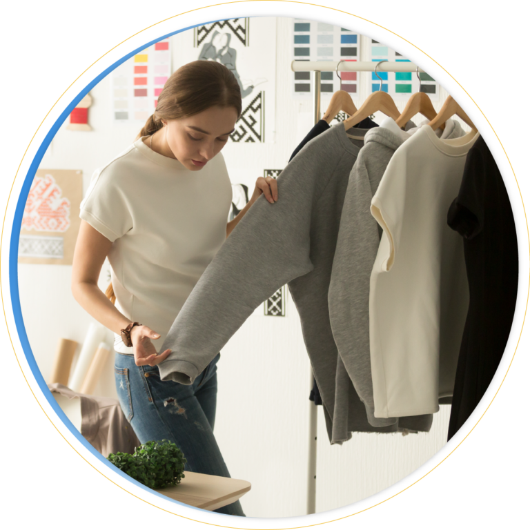 A woman examines a row of sweatshirts on hangers, focusing on a gray one. She is standing in a bright room with color swatches and design patterns in the background, exploring the product assortment.