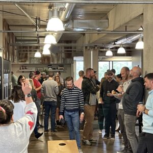 A group of people socialize in a modern office space with exposed ceilings and concrete pillars. Some are chatting, holding drinks or snacks, while others play cornhole, fostering community impact in a casual and lively atmosphere.
