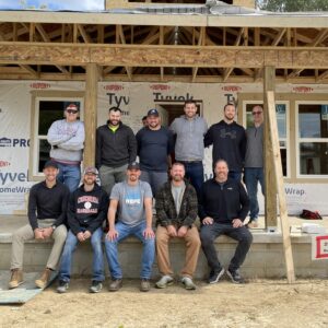 A group of eleven people, some standing and some sitting, pose in front of a partially constructed house with Tyvek wrap and exposed wood beams, showcasing their community impact. They are outdoors on a concrete porch, smiling at the camera.