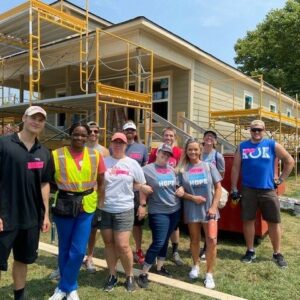 A group of volunteers poses and smiles in front of a house under construction, surrounded by scaffolding. Most wear matching shirts and some have safety vests, showing teamwork and community impact on a sunny day.