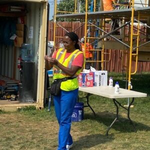 A woman in a safety vest and glasses stands on grass, looking at her phone. Behind her, workers in safety gear are on scaffolding as part of a project with strong community impact. A table with drinks and supplies is nearby amid construction materials.