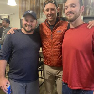 Three men smiling and posing together indoors, reflecting community impact. One wears a blue shirt and hat, holding a drink can; the middle man sports a red vest and cap; the third wears a red shirt. Others and industrial decor are visible in the background.