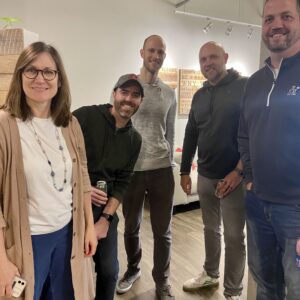 Five adults, four men and one woman, stand indoors on a wooden floor, smiling at the camera. Casually dressed, they appear to be at a social gathering or office event focused on fostering community impact.