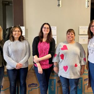 Five women stand smiling in front of a wall with signs and stacked Lowe’s boxes, highlighting their community impact. Casually dressed, they appear to be indoors at an event or volunteer activity.