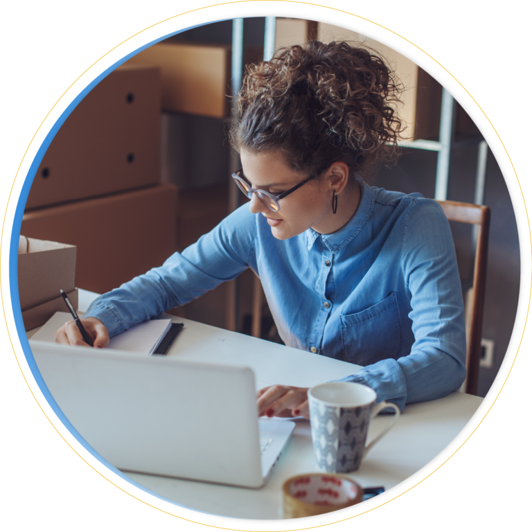 A woman with curly hair and glasses writes in a notebook at her desk, possibly brainstorming merchandise planning solutions, with a laptop, coffee mug, and cardboard boxes in the background. She is wearing a blue button-down shirt.