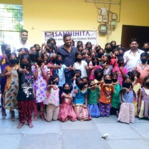 A group of men and many girls, some wearing black face masks, pose together inside a building. A banner behind them reads SANJIHITA, indicating a women and girl children society. The cheerful mood highlights their positive community impact.