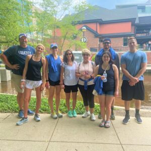 Eight people stand outdoors on a path by a stream, smiling at the camera. Dressed in casual summer clothes and holding water bottles, they gather to celebrate their community impact, with trees and a building in the background.