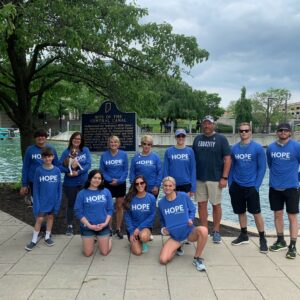 A group of eleven people in matching blue HOPE shirts stand and kneel by a canal, smiling under a tree near a historical marker and stone bridge on a cloudy day, showcasing their community impact.
