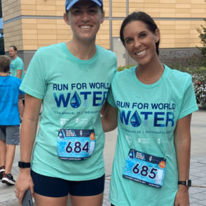 Two women wearing Run for World Water t-shirts and race bibs (684 and 685) smile outdoors at a running event, highlighting community impact, with other participants and a modern building in the background.