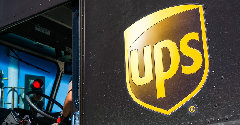 Close-up of a UPS delivery truck showing the yellow UPS shield logo on the black side panel, with part of the drivers arm visible inside the cab.