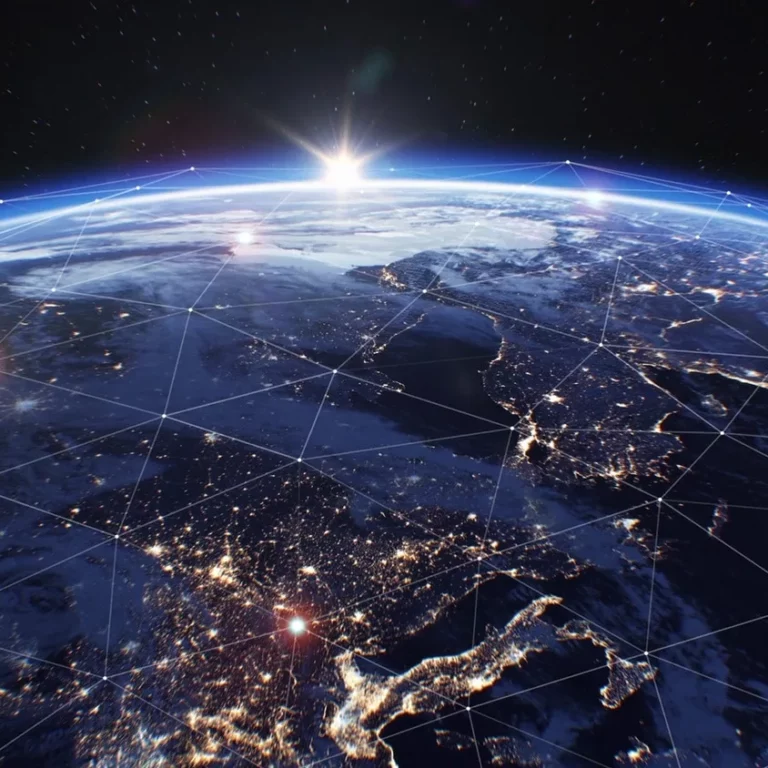 View of Earth from space at sunrise, with illuminated cities and network lines showing global connectivity. The horizon glows with sunlight, and the planets surface features continents and scattered clouds.