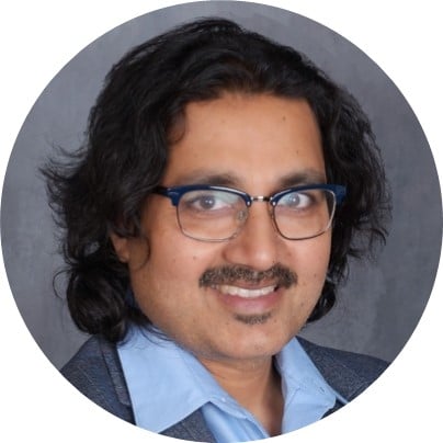 A person with wavy dark hair, glasses, and a mustache, wearing a blue collared shirt and a gray blazer, smiling against a gray background.