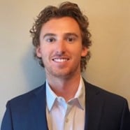A young man with wavy brown hair wearing a dark suit jacket and a white dress shirt, smiling in front of a plain beige background.