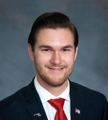 A man in a dark suit, white shirt, and red tie is smiling at the camera. He has short, neatly styled brown hair and a trimmed beard. An American flag pin is on his lapel, and the background is plain gray.