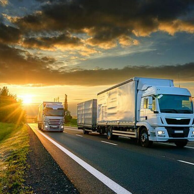Two large trucks drive on a highway through green fields under a dramatic, cloudy sky at sunrise or sunset, with sunlight streaming onto the road.