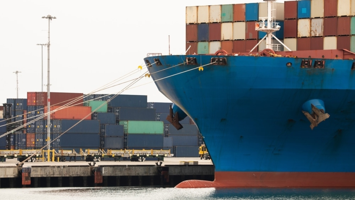 A large blue cargo ship docked at a port, with colorful shipping containers stacked both on the vessel and on the dock in the background.