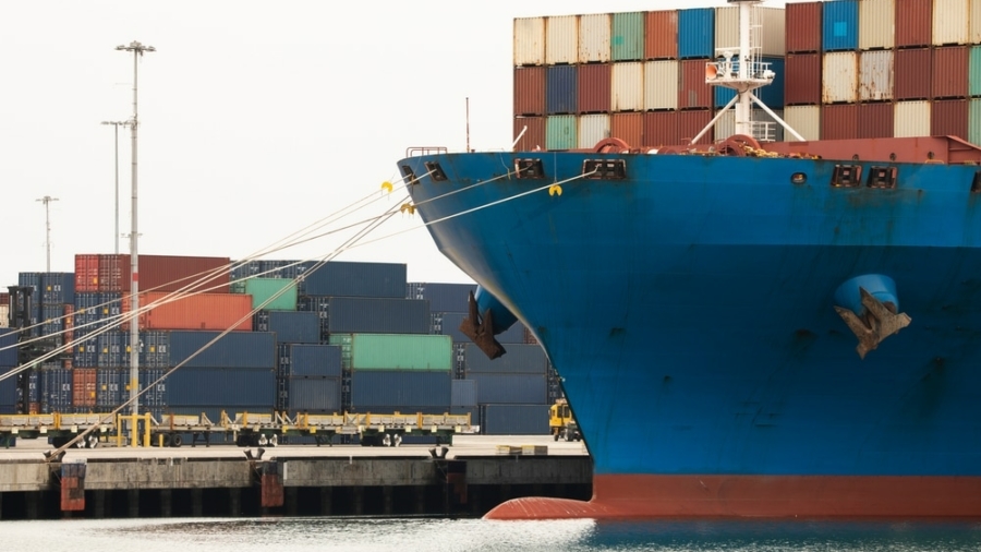 A large blue cargo ship docked at a port, with colorful shipping containers stacked both on the vessel and on the dock in the background.