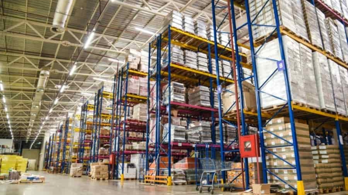 Large warehouse interior with tall blue and yellow shelving racks stacked with various pallets and boxes, organized in rows. The space is brightly lit with high ceilings and an orderly layout.