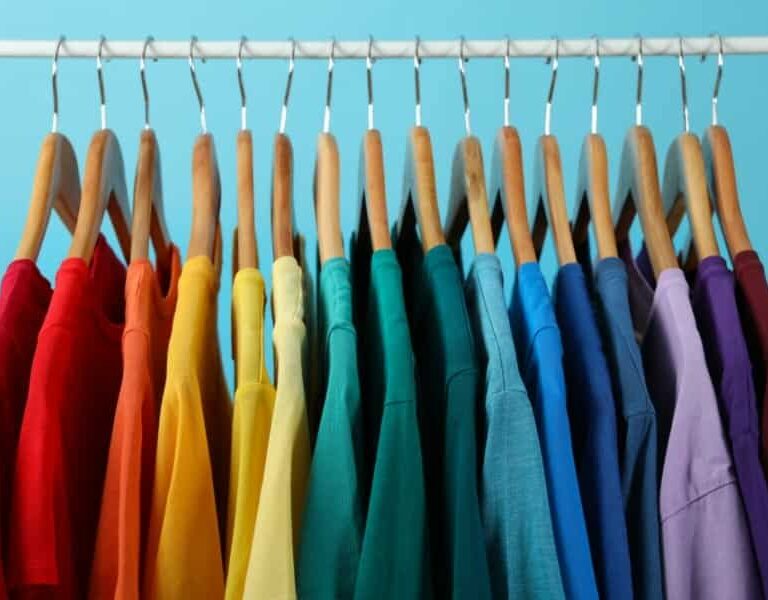 A row of colorful t-shirts hanging on wooden hangers, arranged in a gradient rainbow order, against a light blue background.