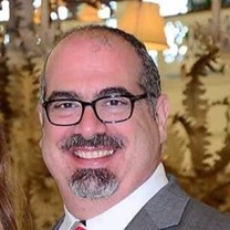 A middle-aged man with glasses, a short gray beard, and a mustache is smiling in a gray suit and tie, possibly discussing the bullwhip effect, with lamps and plants blurred in the background.