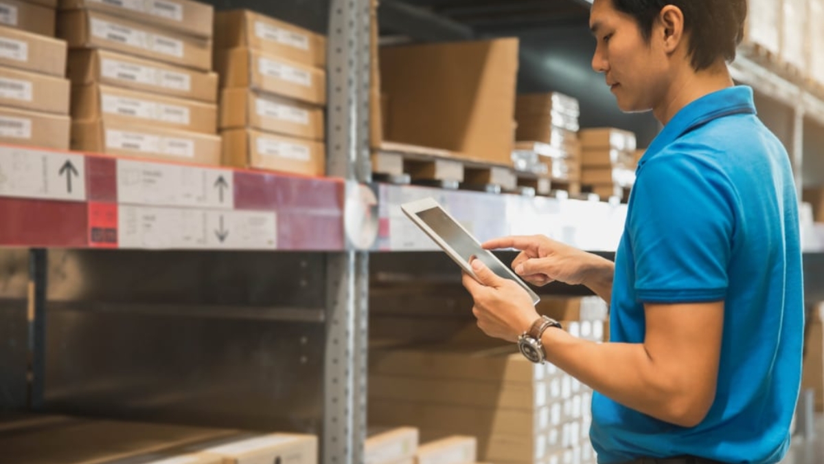 A man in a blue polo shirt uses a tablet while standing in a warehouse aisle of micro-fulfillment centers, surrounded by shelves stacked with labeled cardboard boxes.