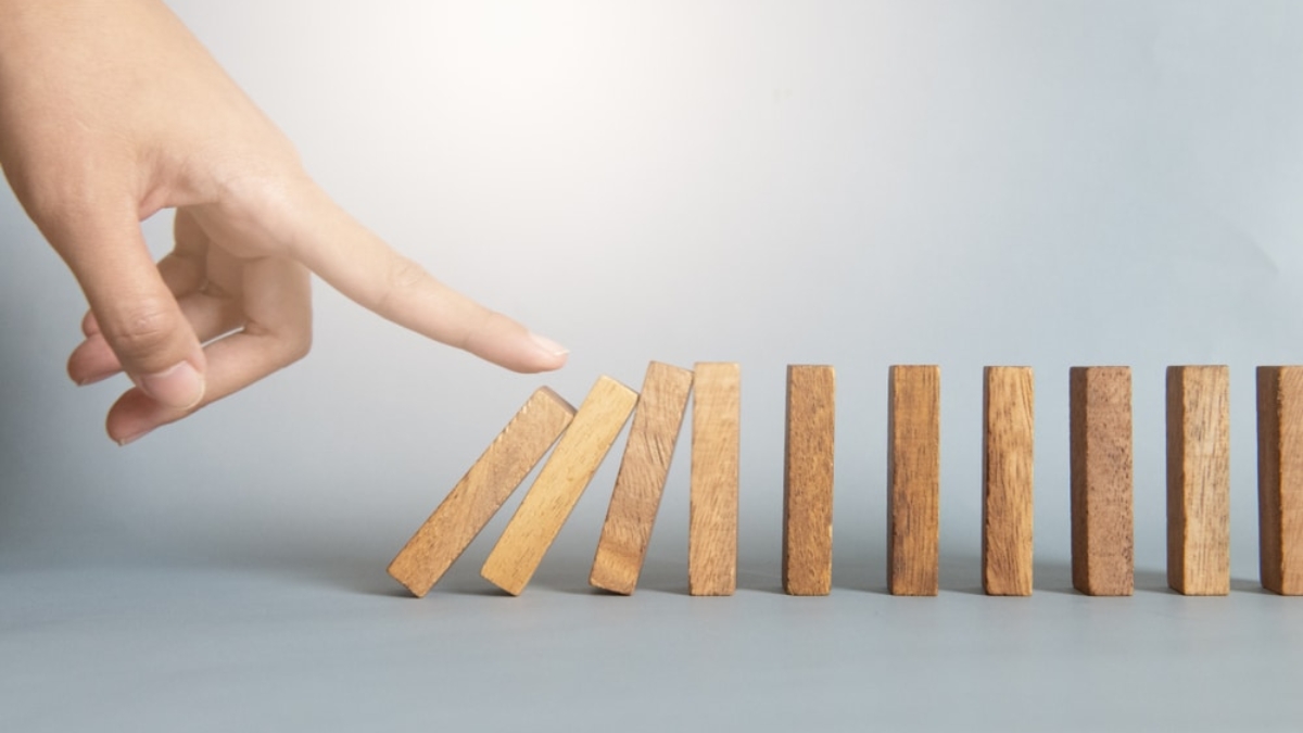 A hand is about to tip over the first of several upright wooden dominoes, illustrating a bullwhip effect chain reaction. The background is plain and light-colored.