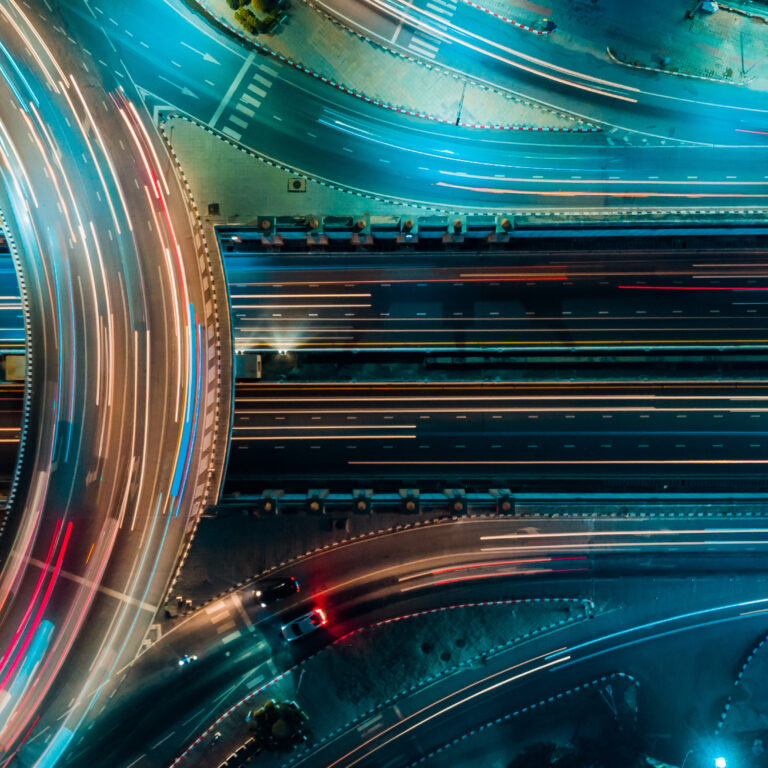 Aerial view of a brightly lit highway interchange at night, showing light trails from moving vehicles and curved roads intersecting with multiple lanes, surrounded by illuminated urban areas.