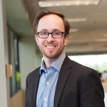 A man with glasses, short brown hair, and a beard, wearing a dark blazer and checked shirt, smiles while standing in a modern office hallway with large windows and bright lighting.