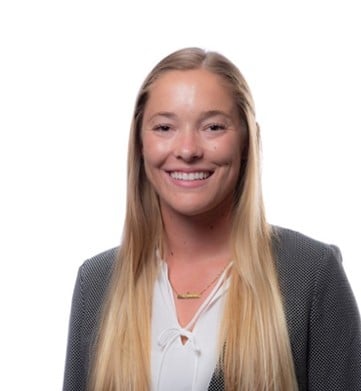 A woman with long blonde hair wearing a grey blazer over a white blouse smiles at the camera against a plain white background.