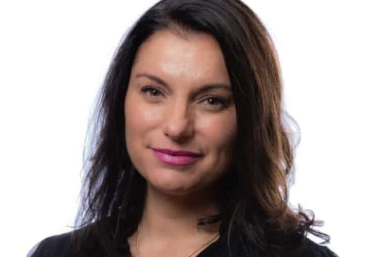 A woman with long dark hair and a slight smile, wearing a black top, is pictured against a plain white background.