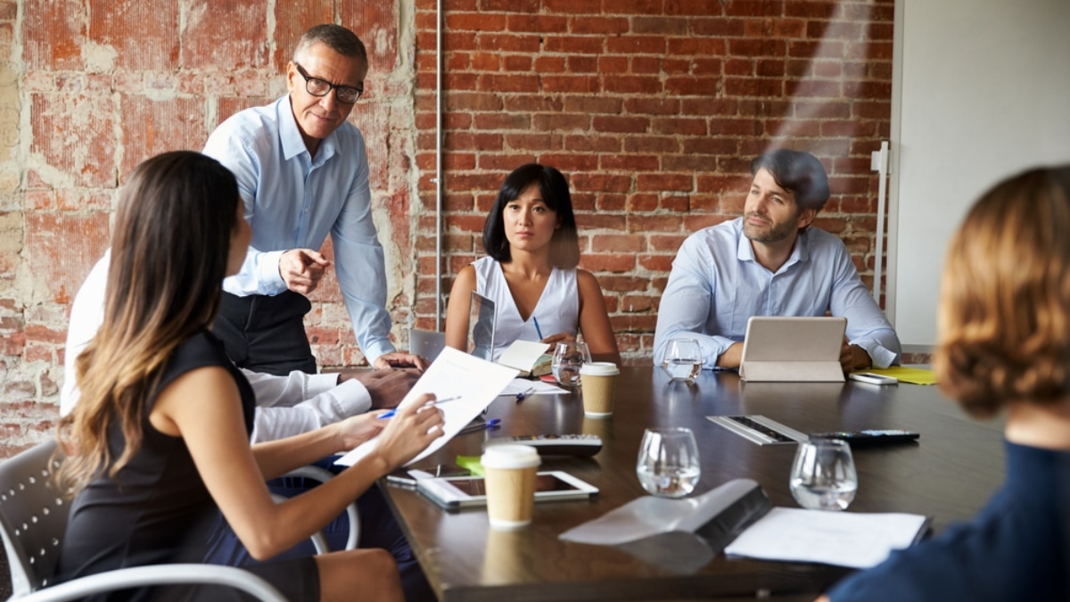 A group of five people sits around a table in a modern office with exposed brick walls, engaged in a meeting. One man is standing and speaking, while others listen and take notes. Laptops, papers, and drinks are on the table.