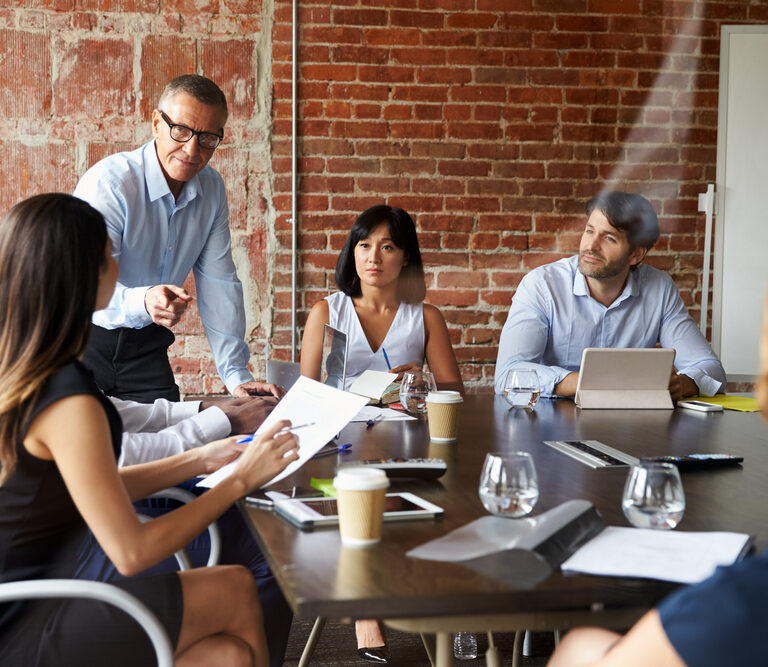 A group of five people sits around a table in a modern office with exposed brick walls, engaged in a meeting. One man is standing and speaking, while others listen and take notes. Laptops, papers, and drinks are on the table.