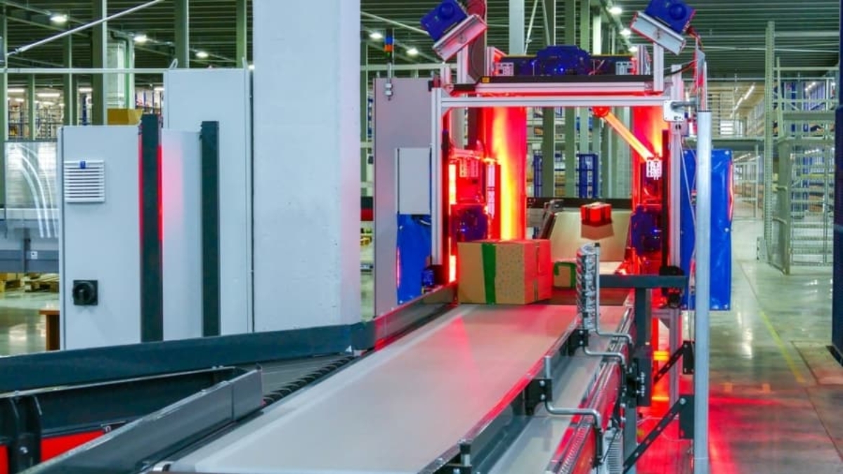 A conveyor belt in a warehouse moves packages through a scanning machine with red lights, surrounded by metal structures and shelving in an industrial setting.