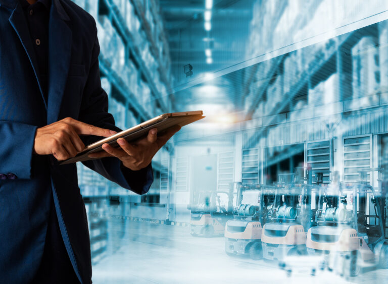 A person in a blue suit uses a tablet, with a modern warehouse filled with shelves and forklifts in the background, symbolizing logistics, inventory management, or supply chain planning operations.