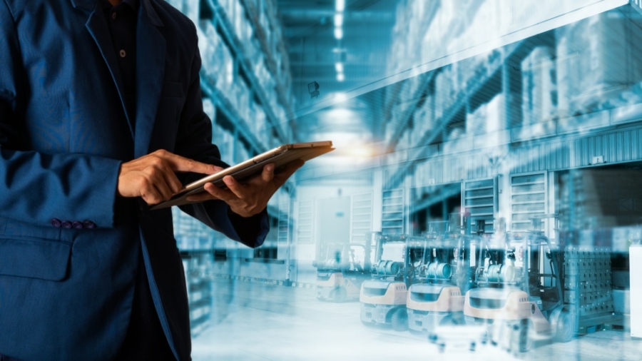 A person in a blue suit uses a tablet, with a modern warehouse filled with shelves and forklifts in the background, symbolizing logistics, inventory management, or supply chain planning operations.