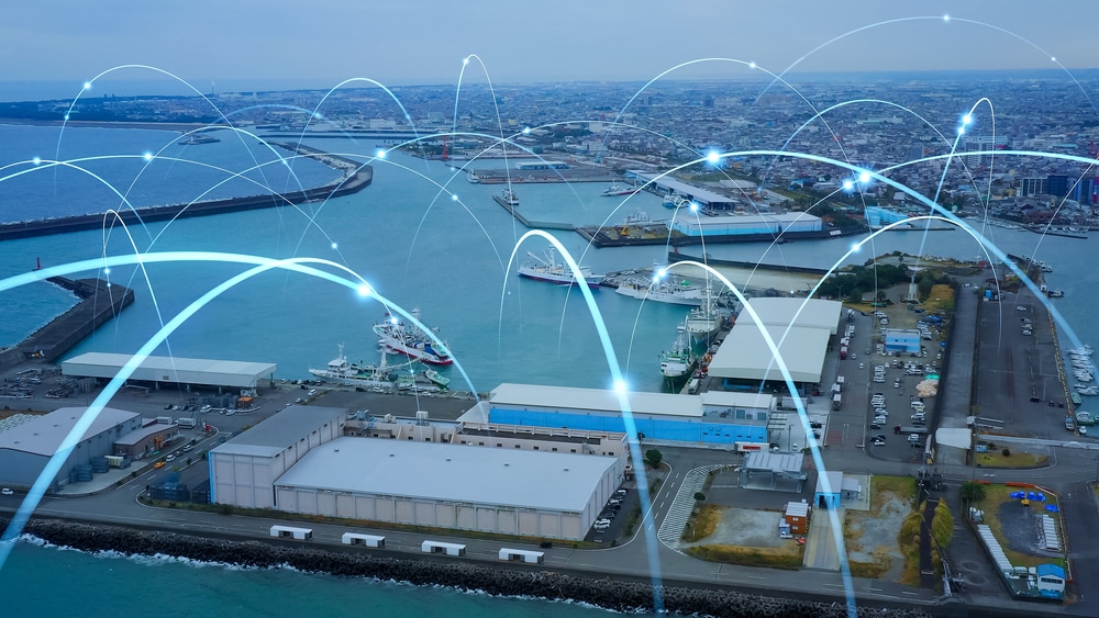 Aerial view of a busy port with warehouses, ships, and blue water. Digital lines and glowing points are overlaid, symbolizing a responsive supply chain with seamless network connectivity and efficient data transfer across the port.