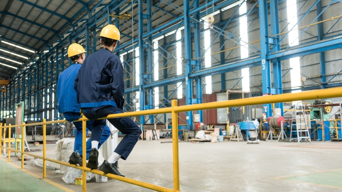 Two workers in blue uniforms and yellow hard hats sit on a yellow metal railing inside a spacious industrial warehouse with high ceilings and blue structural beams.