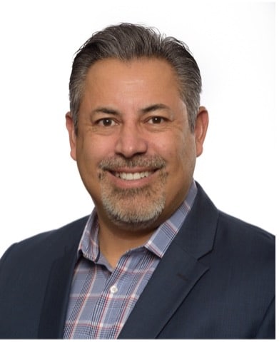 Smiling man with short gray hair, a beard, and mustache, wearing a dark blazer over a plaid shirt, posed against a plain white background.
