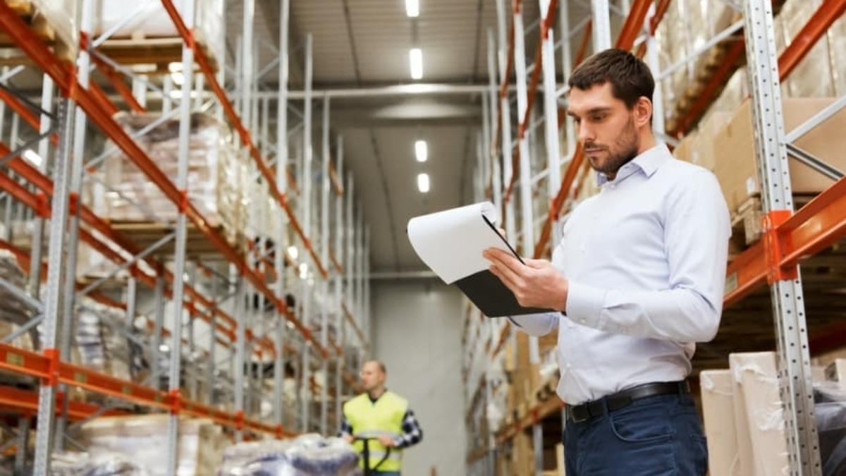 A man in a white shirt holds a clipboard and checks inventory in a large warehouse with tall shelves, while another worker in a safety vest walks in the background.