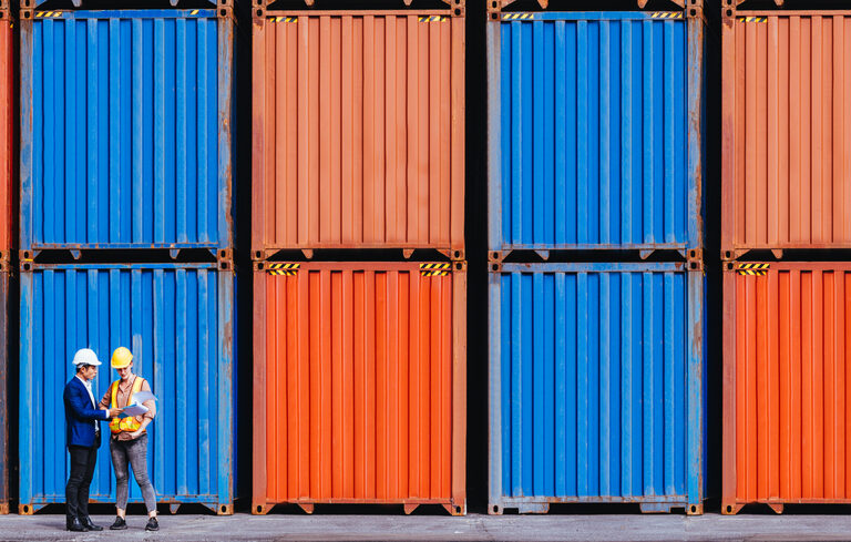 Two people wearing helmets stand in front of stacked orange and blue shipping containers, appearing to have a discussion at a shipping yard or port.