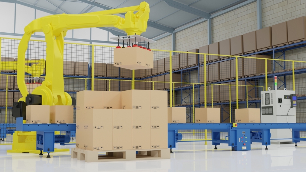 A yellow robotic arm performs automated palletization, moving cardboard boxes onto a conveyor belt in a clean, brightly lit warehouse, with shelves of additional boxes in the background.