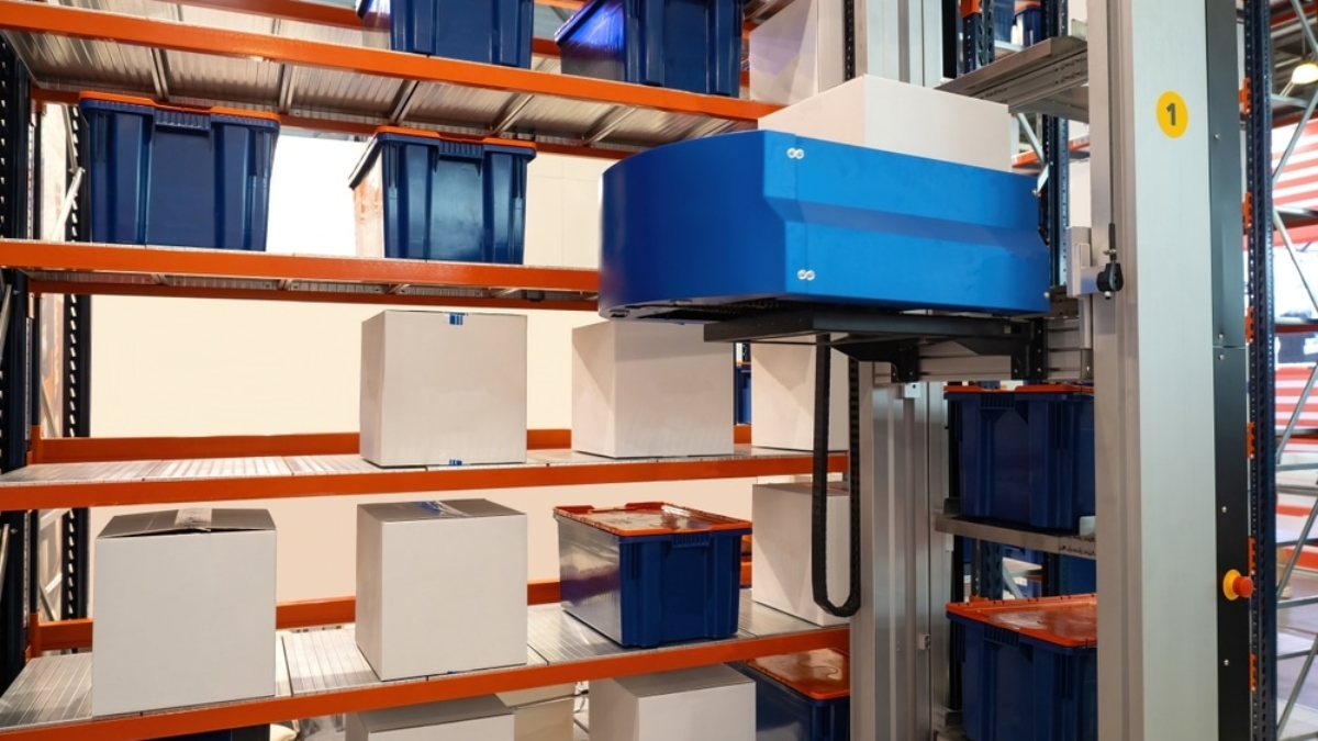 Automated warehouse system with robotic arm stacking and moving blue bins and white boxes on metal shelving units.