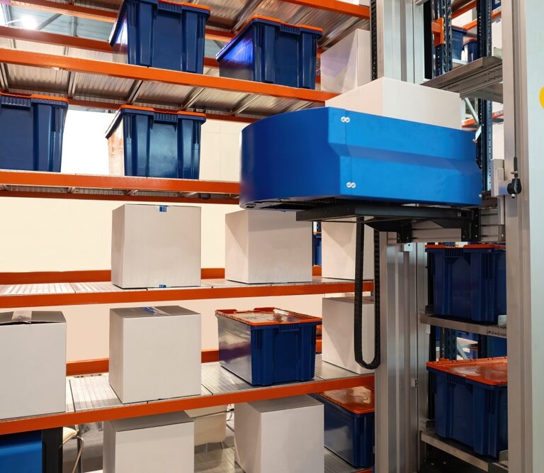 Automated warehouse system with robotic arm stacking and moving blue bins and white boxes on metal shelving units.