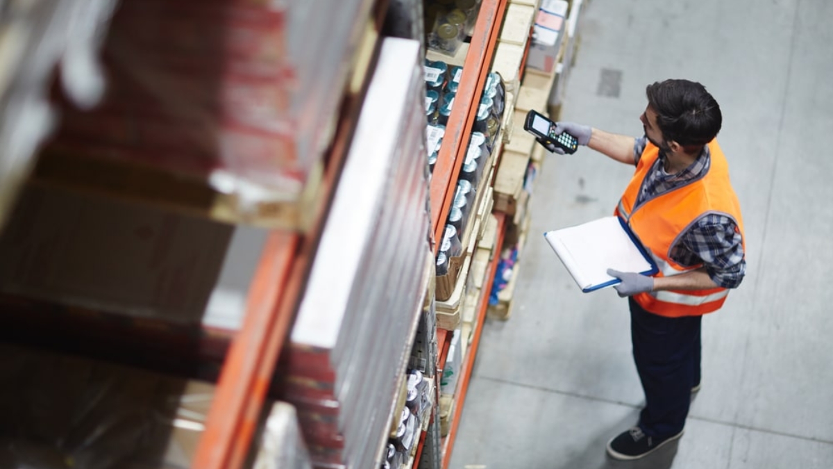 A warehouse worker in an orange safety vest scans items on a high shelf with a handheld device while holding a clipboard, standing in an industrial storage area.