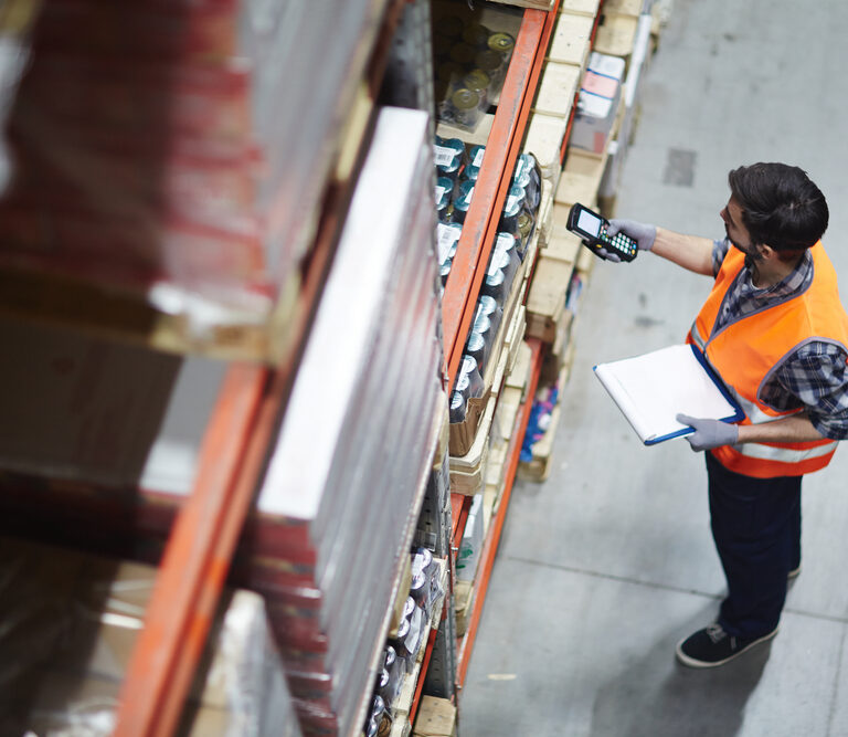 A warehouse worker in an orange safety vest scans items on a high shelf with a handheld device while holding a clipboard, standing in an industrial storage area.