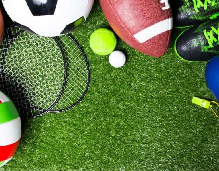Various sports equipment, including soccer ball, football, volleyball, tennis rackets, tennis ball, golf ball, green sports shoes, blue boxing glove, and a yellow whistle, arranged on green artificial grass.