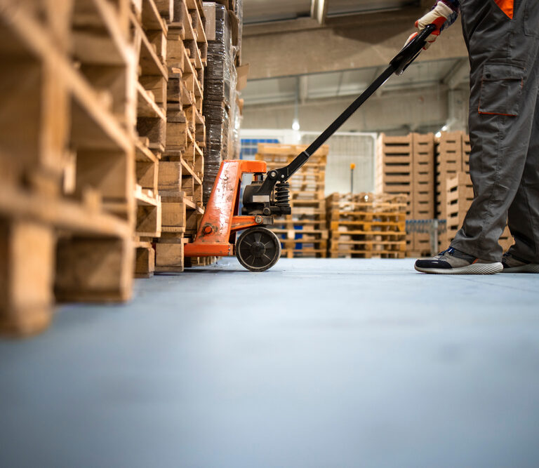 A worker in gray pants and sneakers operates an orange pallet jack, moving wooden pallets inside a warehouse with stacks of pallets in the background.